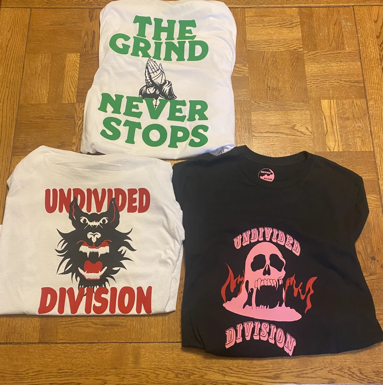 Original Undivided Division white Wolf, pink skull and The Grind Never Stops tees laid out together